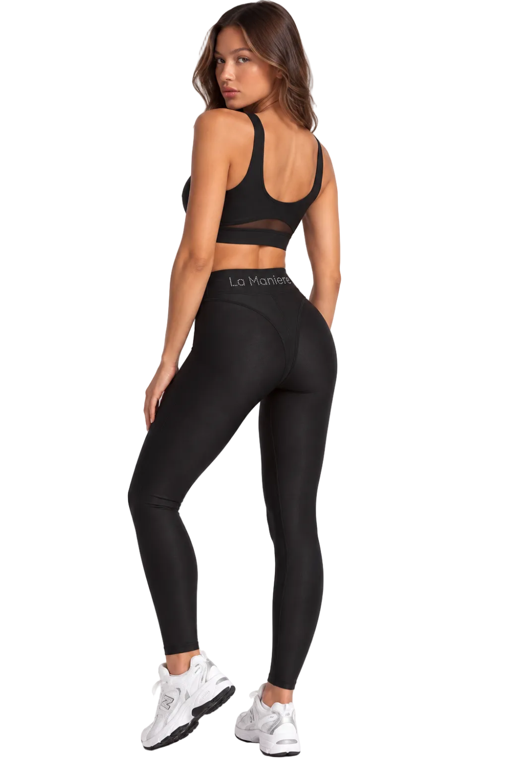A woman modeling black athletic wear, featuring a black sports bra with a sheer back panel and black high-waisted leggings with 'La Marianne' printed on the waistband, paired with white sneakers against a plain white studio background.