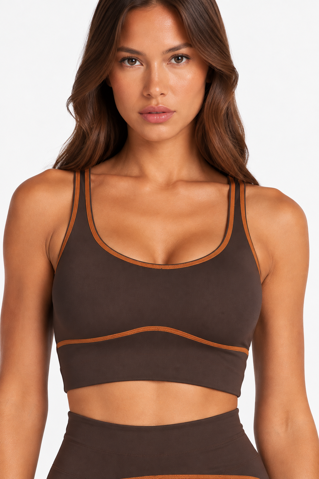 A woman wearing a dark brown sports bra with thin tan contrast trim and a matching high-waisted brown bottom, shown against a plain light background.